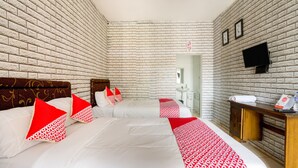 A hotel room with a brick-patterned wall, a bed with white bedding and red pillows, a bedside table with a clock, and a small desk with a telephone and a cup.