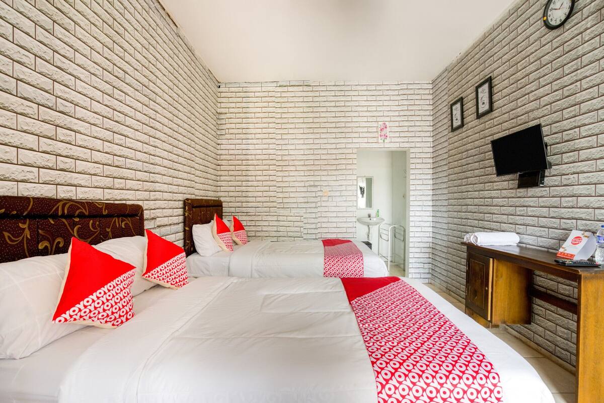 A hotel room with a brick-patterned wall, a bed with white bedding and red pillows, a bedside table with a clock, and a small desk with a telephone and a cup.