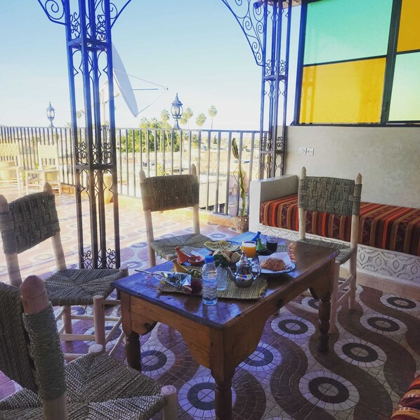 A terrace with wicker furniture, a table set with food and drinks, and a view of the cityscape. - RIAD MALAYINE (Marrakech)