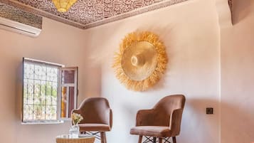 Une pièce avec un plafond orné de motifs, deux chaises, une table sur laquelle se trouve un vase, et une fenêtre agrémentée d’une grille décorative.