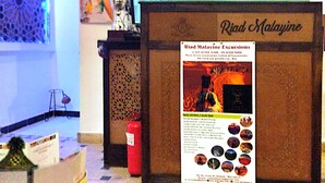 A reception desk with a sign for Riad Malajine Excursions, a small table with a decorative item, and a patterned carpet. - RIAD MALAYINE (Marrakech)