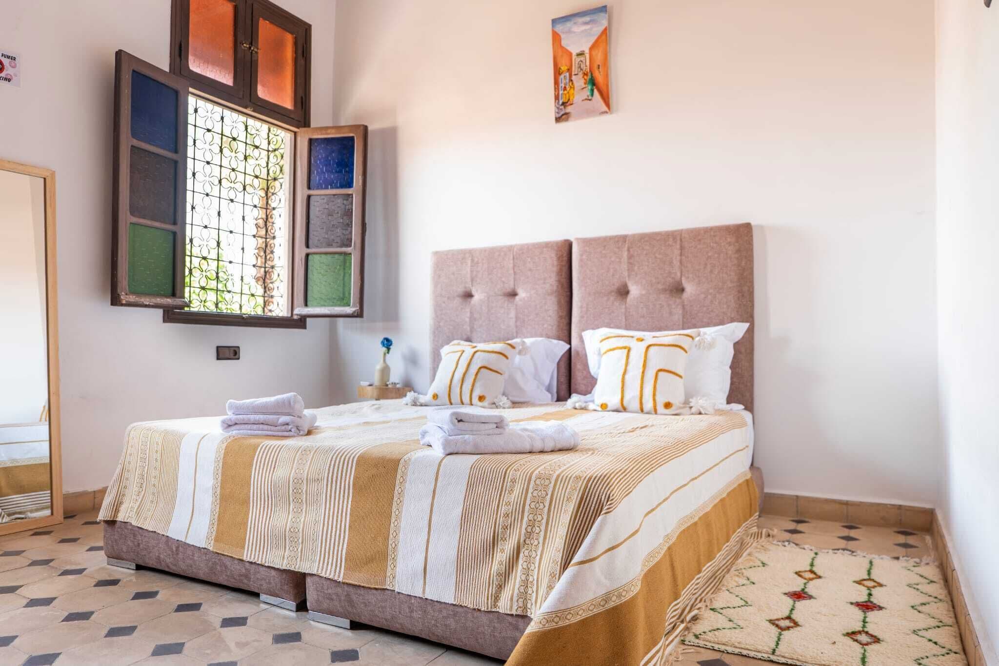 Una cama bien tendida con una colcha a rayas, dos almohadas con diseño geométrico y una ventana con celosía decorativa.