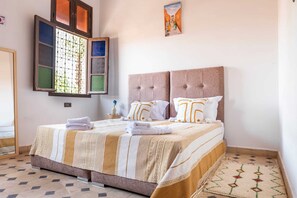 Una cama bien tendida con una colcha a rayas, dos almohadas con diseño geométrico y una ventana con celosía decorativa.