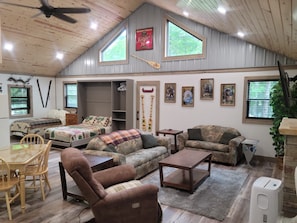 Ein gemütliches Wohnzimmer mit hoher Decke, Holzbalken und großen Fenstern. Es gibt ein Sofa, einen Sessel und einen kleinen Tisch.