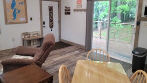 A cozy room with a wooden ceiling, a map on the wall, a door with a decorative hanging, a sign offering free beer, a table with chairs, and a view of a green outdoor area through a sliding door.