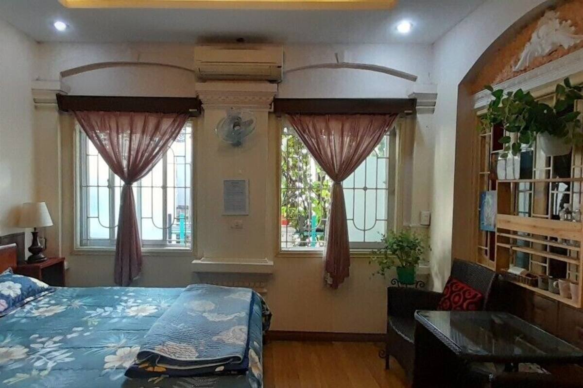 A room with a bed, a desk, a chair, a window with curtains, a fan, and a shelf with items.