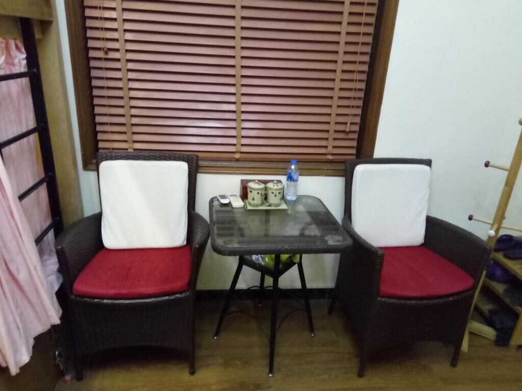 A small room with two wicker chairs with red cushions, a glass table with a few items, and a window with blinds.