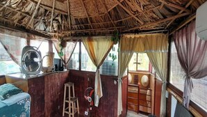 A room with a thatched roof, wooden walls, a fan, a small table, a chair, and a window with curtains. - Green Homestay Ha Noi (Hanoi)