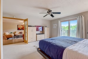 Ein Schlafzimmer mit Bett, Deckenventilator, Fernseher, Schreibtisch und einem Balkon mit Vorhängen.