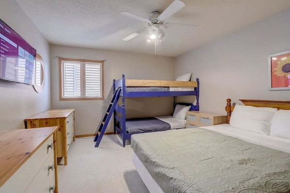 Une chambre avec des lits superposés, un bureau, une chaise, un ventilateur de plafond et une fenêtre avec des stores.