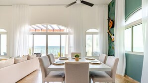 A dining area with a white table set for four, surrounded by beige chairs, with a view of the sea through large windows. - Refurbished BeachFront Penthouse - Music themed! (Cabarete)