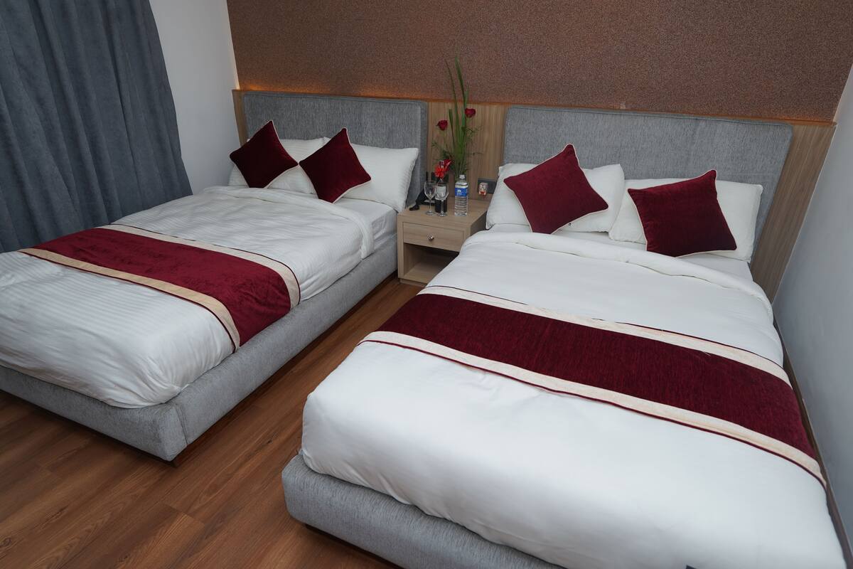 A hotel room with two beds, each with white bedding and red accents, a wooden floor, and a nightstand with a vase of flowers.
