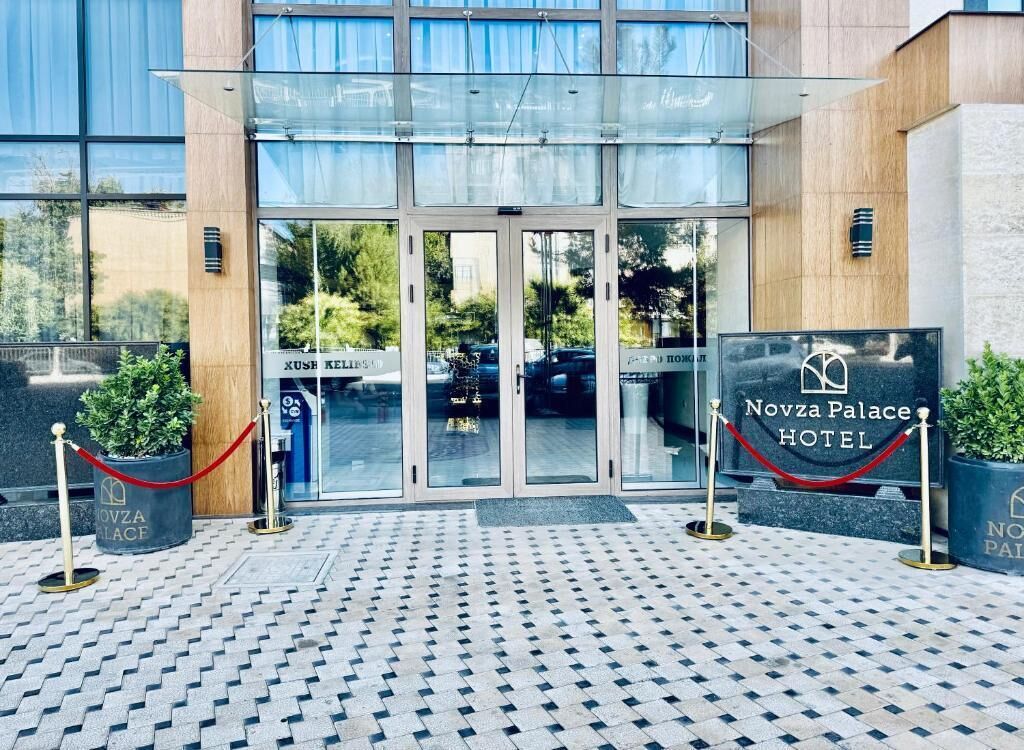 The entrance to Novza Palace Hotel with a glass door, a tiled floor, and potted plants.