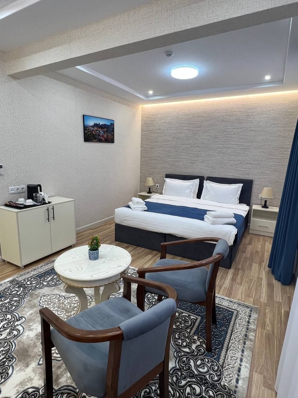 A hotel room with a bed, bedside tables, a desk with a chair, a small round table with a chair, and a decorative rug.