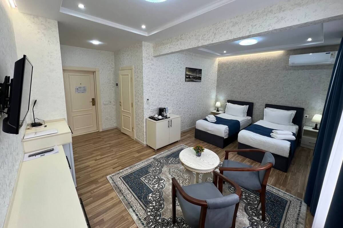 A hotel room with a desk, two beds, a small table with chairs, a TV, and a patterned rug.