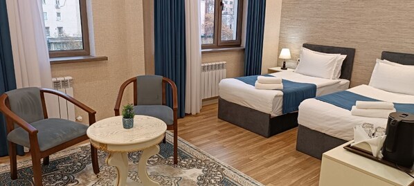Une chambre d’hôtel avec deux lits, une petite table, deux chaises, un radiateur et une fenêtre avec des rideaux.