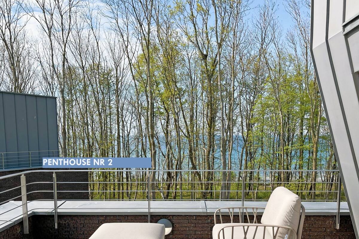 A rooftop terrace with two lounge chairs and a table, overlooking a forest and a body of water.