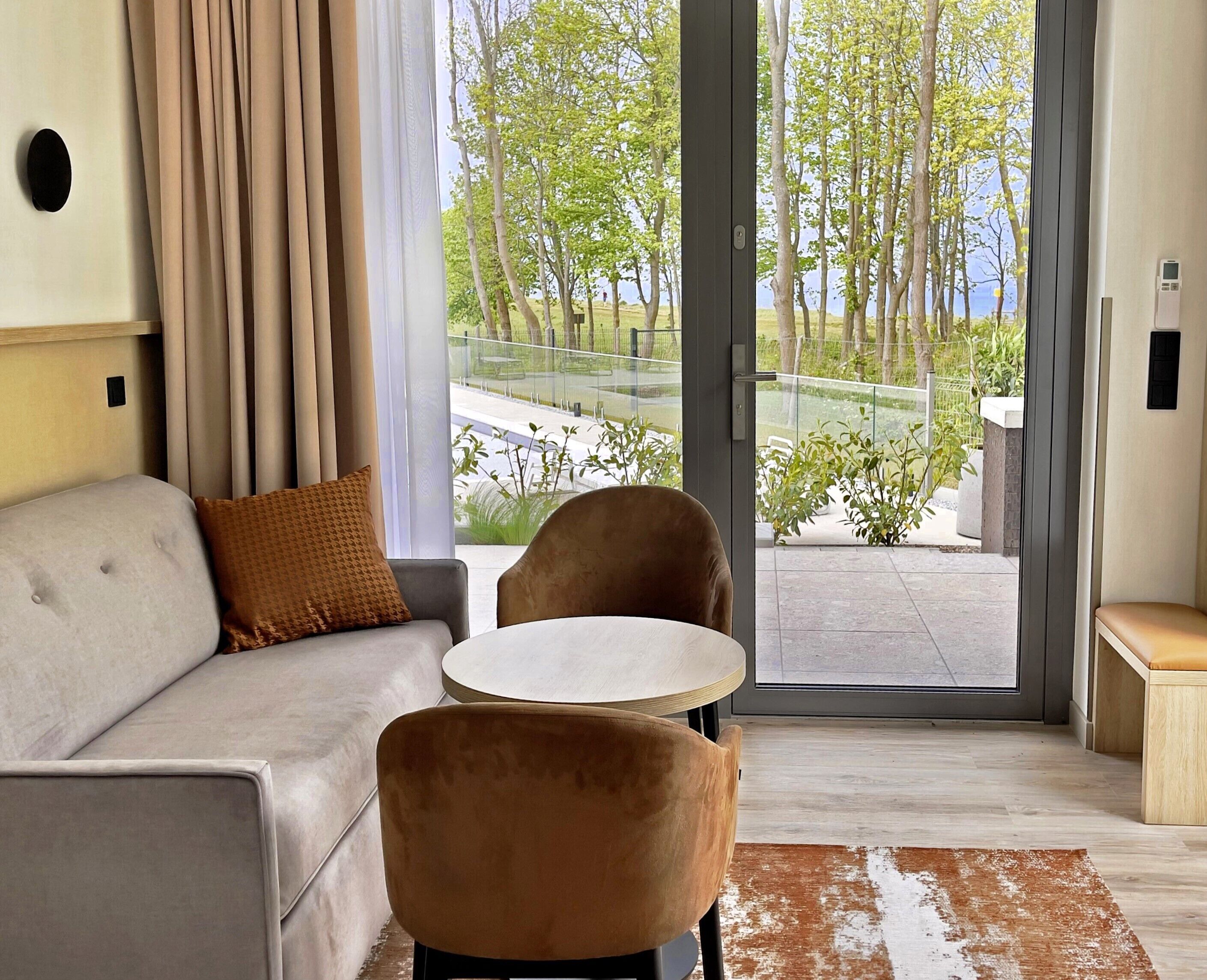 A modern room with a sofa, two chairs, a small table, and a view of a balcony with plants and a railing.