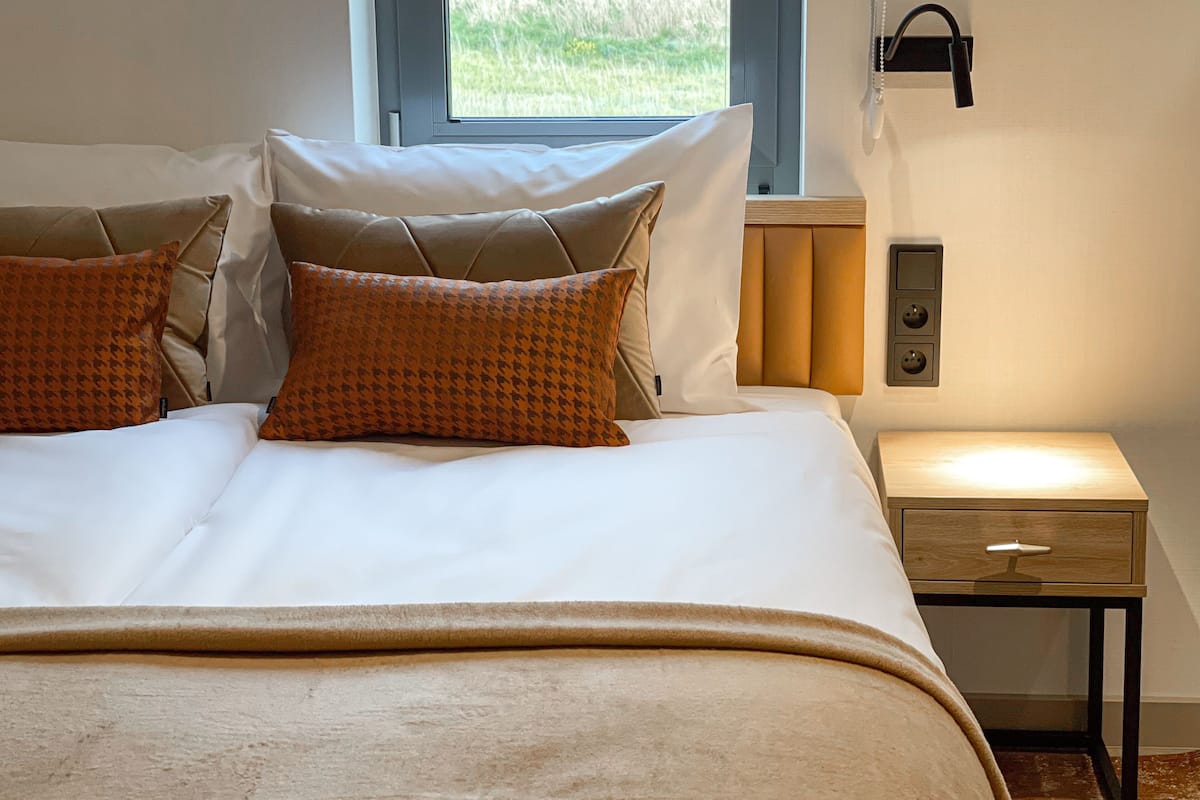 A neatly made bed with a white duvet and two patterned pillows, a bedside table with a lamp, and a window with a view of greenery.