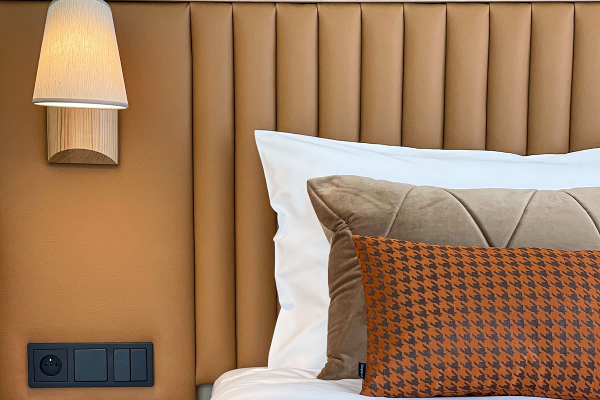 A neatly made bed with a white and brown pillow, a patterned pillow, and a beige blanket. There is a bedside table with a lamp, a power outlet, and a drawer.