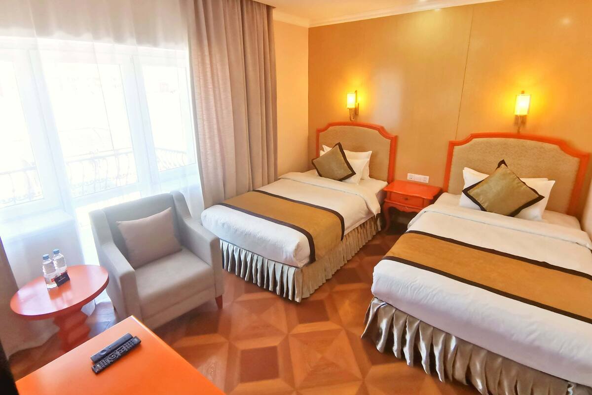 Ein Hotelzimmer mit zwei Betten, einem kleinen Tisch mit einer Fernbedienung, einem Stuhl und einem Fenster mit Vorhängen.