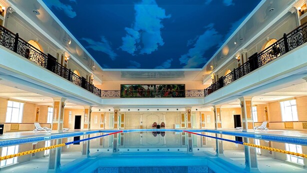A large indoor swimming pool with a ceiling mural of clouds, surrounded by balconies with railings and lighting. - Bayangol Hotel & Resort (Ulaanbaatar)
