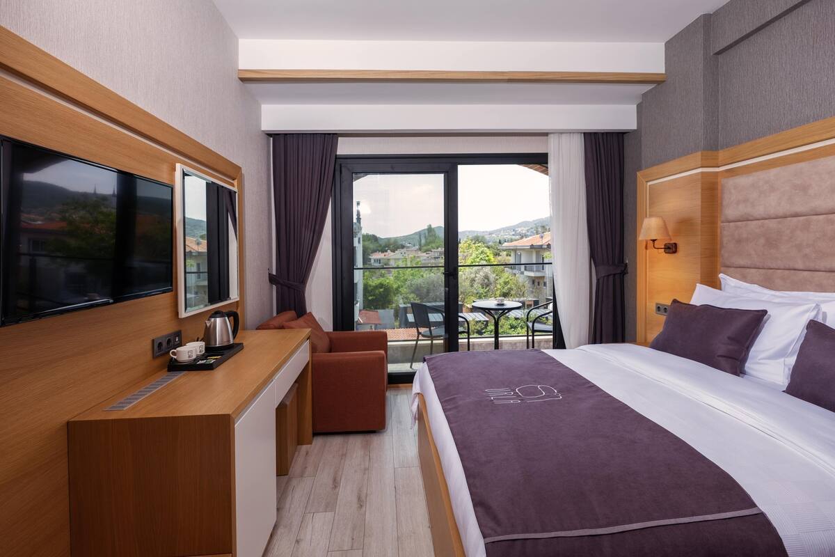 A modern hotel room with a large bed, a desk with a flat-screen TV, a balcony with a table and chairs, and a scenic view of the city.
