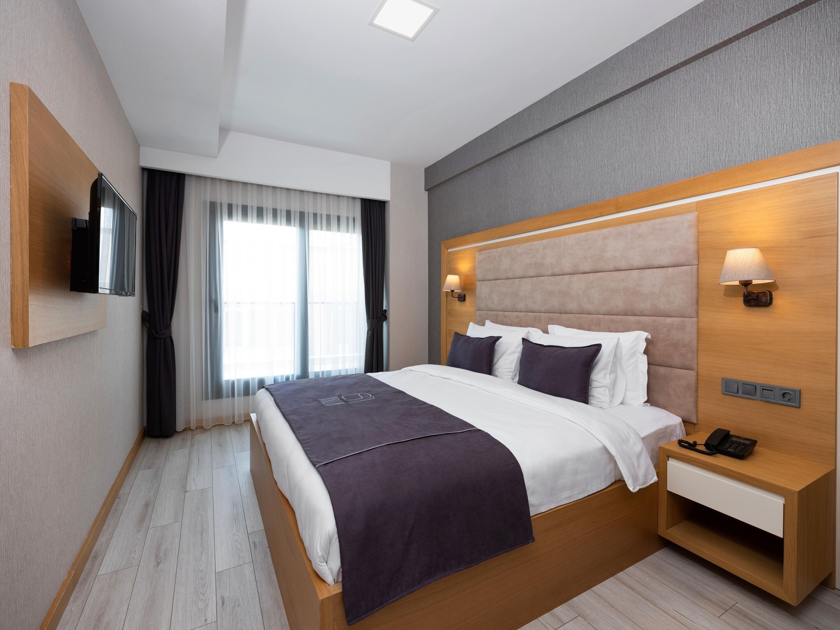 Una habitación de hotel moderna con una cama grande, una mesita de noche con lámpara, un televisor en la pared y una ventana con cortinas.