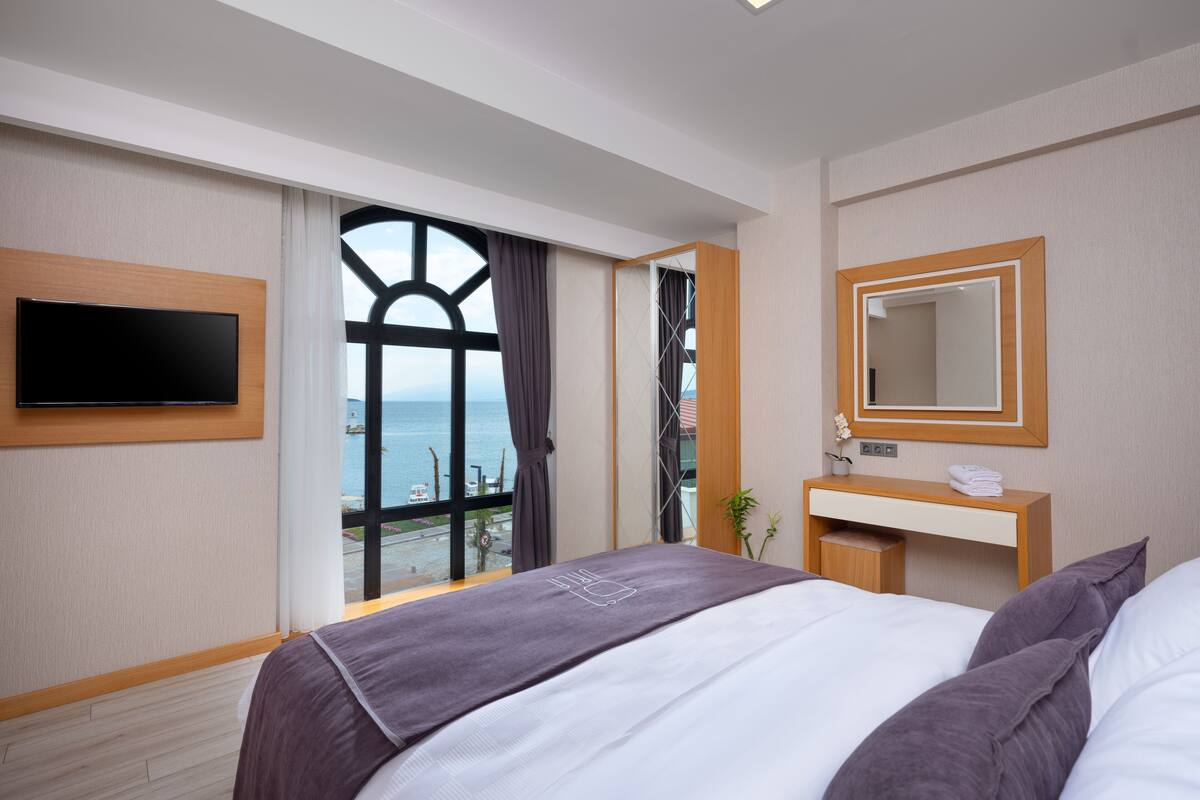 A hotel room with a bed, a TV mounted on the wall, a bedside table with a mirror, and a window with a view of the sea.
