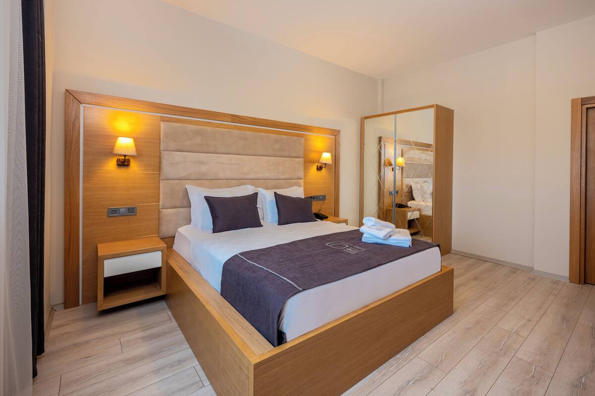 A modern hotel room with a large bed, wooden headboard, bedside tables with lamps, and a mirror.