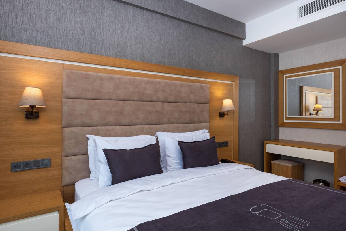 A modern hotel room with a large bed, two bedside lamps, a wooden headboard, a bedside table, and a mirror on the wall.