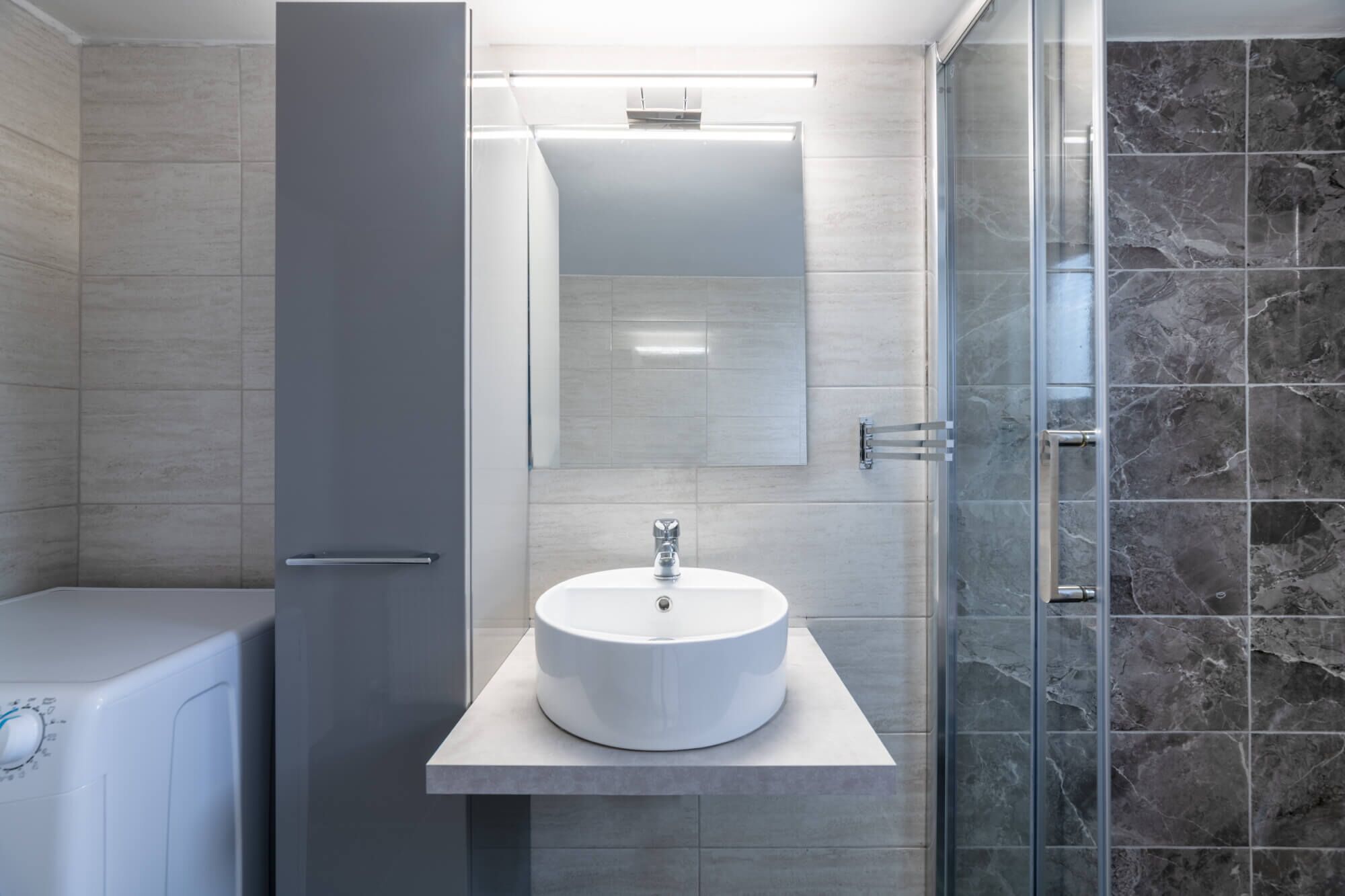 A modern bathroom with a white sink, a mirror, and a shower area with dark tiles.