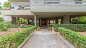 Une allée couverte, dotée d’une porte centrale et bordée de verdure et de plantes en pot, mène à l’entrée d’un bâtiment.