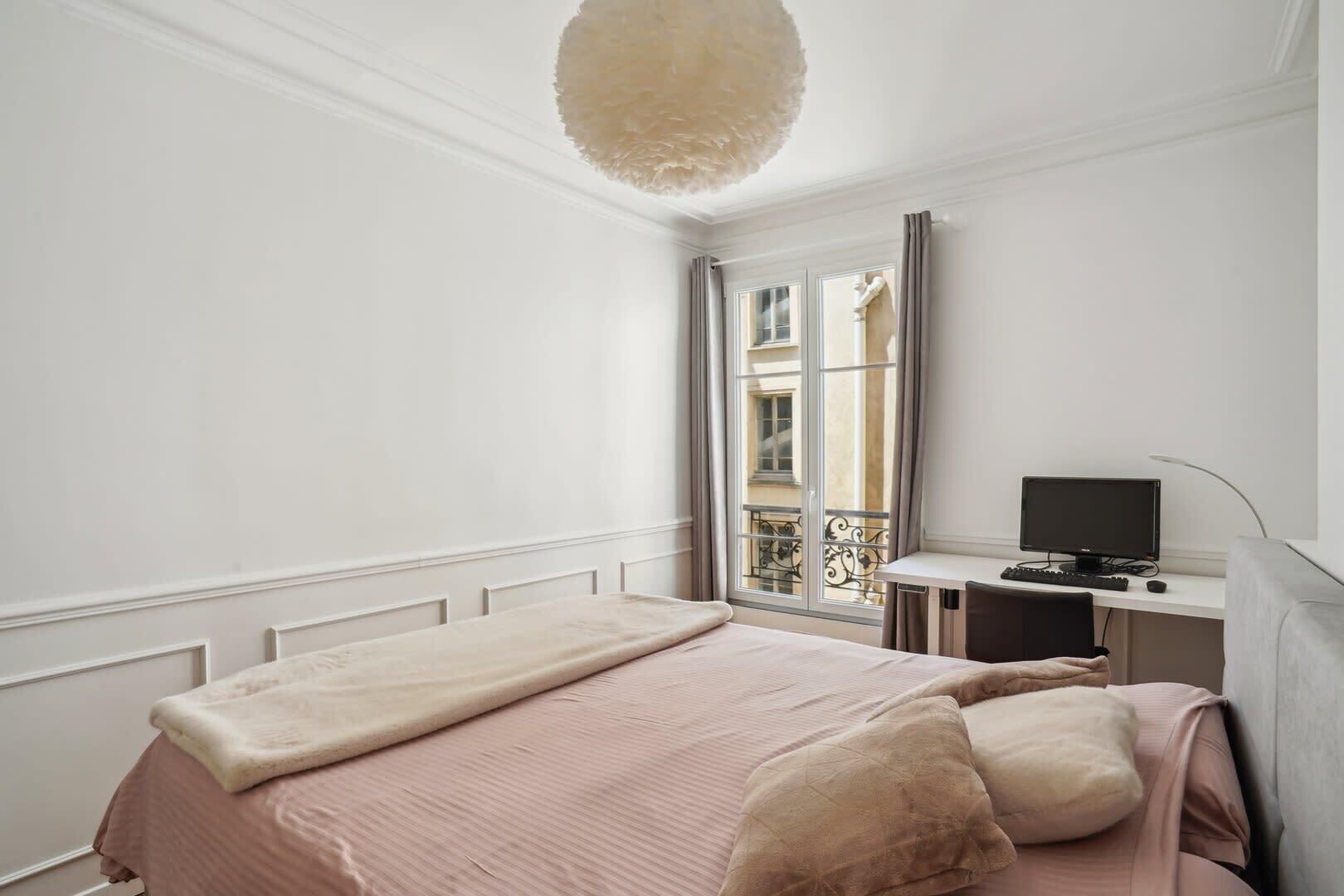 Une chambre à coucher avec un lit, un bureau avec un ordinateur et une fenêtre donnant sur des immeubles.