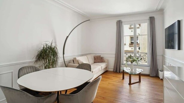 Ett modernt vardagsrum med ett runt matbord, grå klädda stolar, en beige soffa, ett soffbord i glas, en krukväxt och utsikt över en balkong genom ett fönster med grå gardiner.