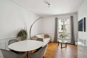 Ein modernes Wohnzimmer mit einem runden Esstisch, grau gepolsterten Sesseln, einem beigen Sofa, einem Glastisch, einer Topfpflanze und Blick auf einen Balkon durch ein Fenster mit grauen Vorhängen.
