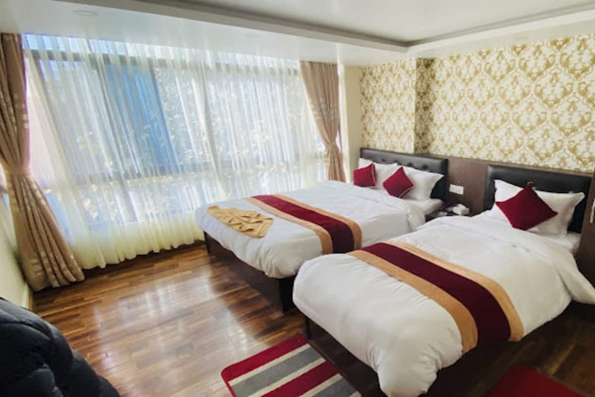 A hotel room with two beds, wooden flooring, a large window with curtains, and patterned wallpaper.