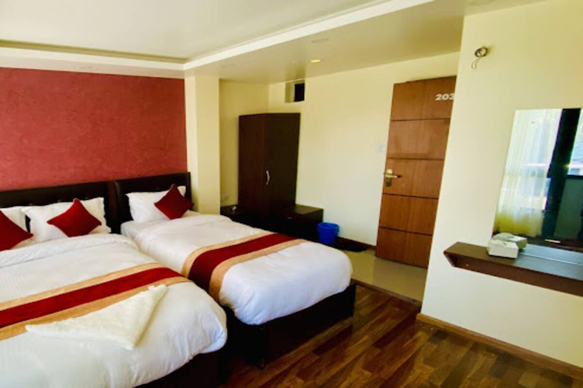 A hotel room with two beds, a red accent wall, a wooden floor, a wardrobe, and a small table with a mirror.