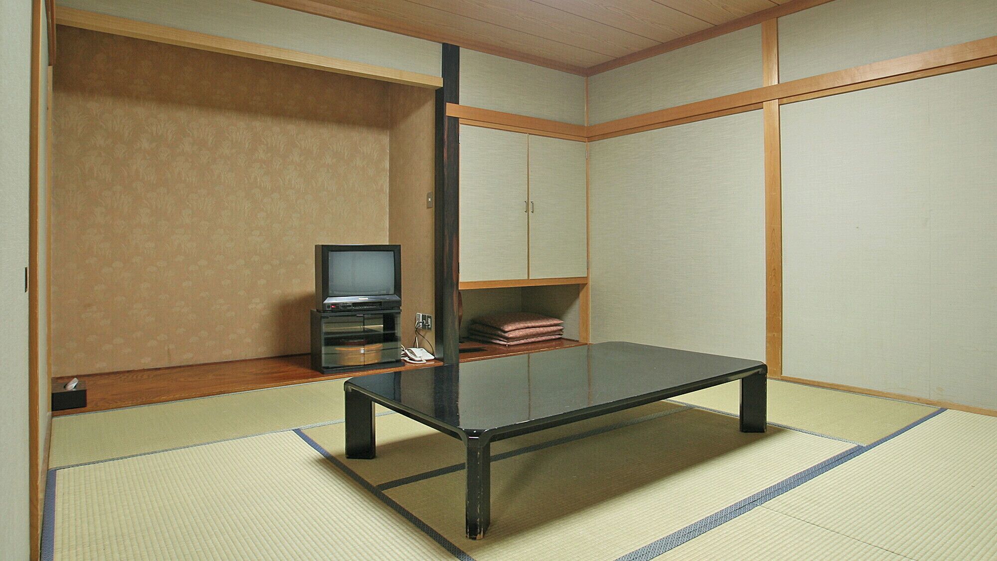 Ein traditioneller japanischer Raum mit Tatami-Boden, einem niedrigen Tisch, einem Fernseher auf einem Schrank und einem Schrank mit zusammengeklappten Gegenständen.