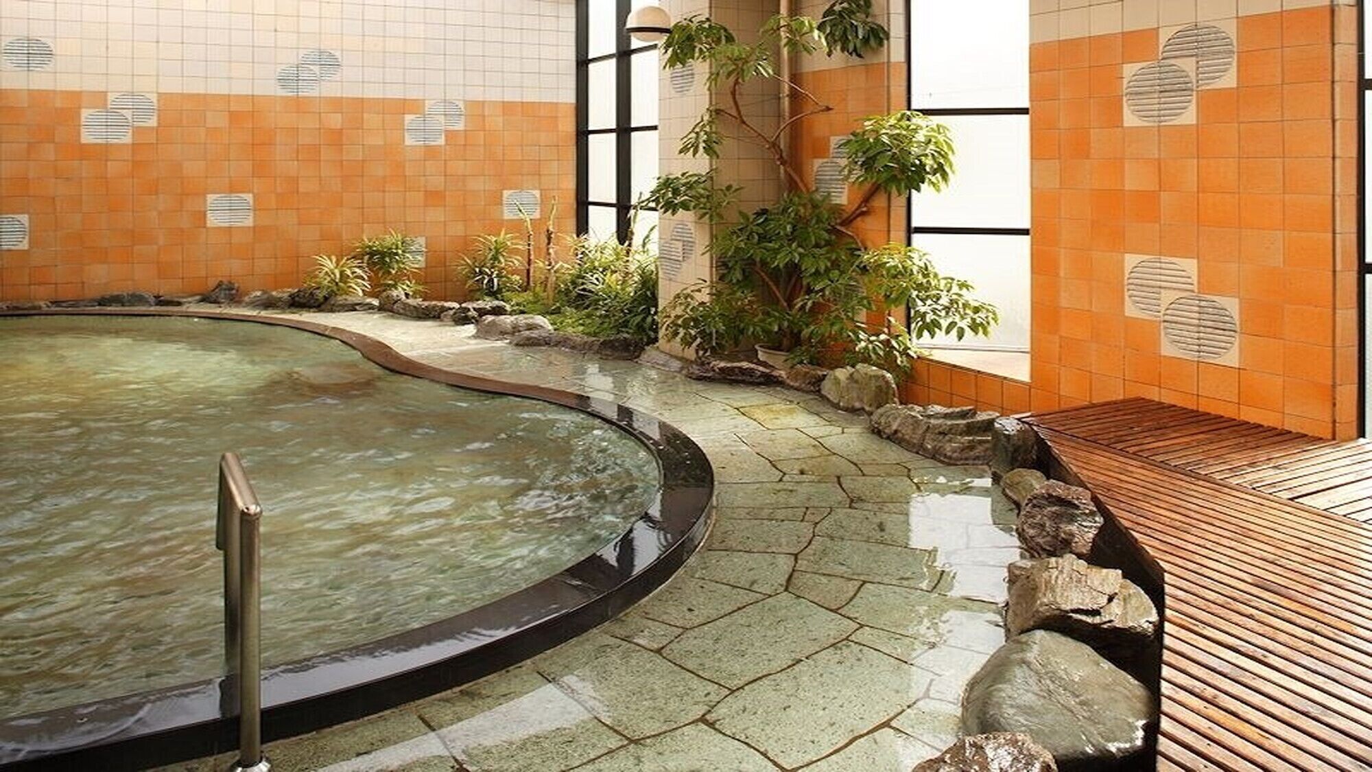 An indoor swimming pool with a wooden deck, surrounded by plants and stone tiles.