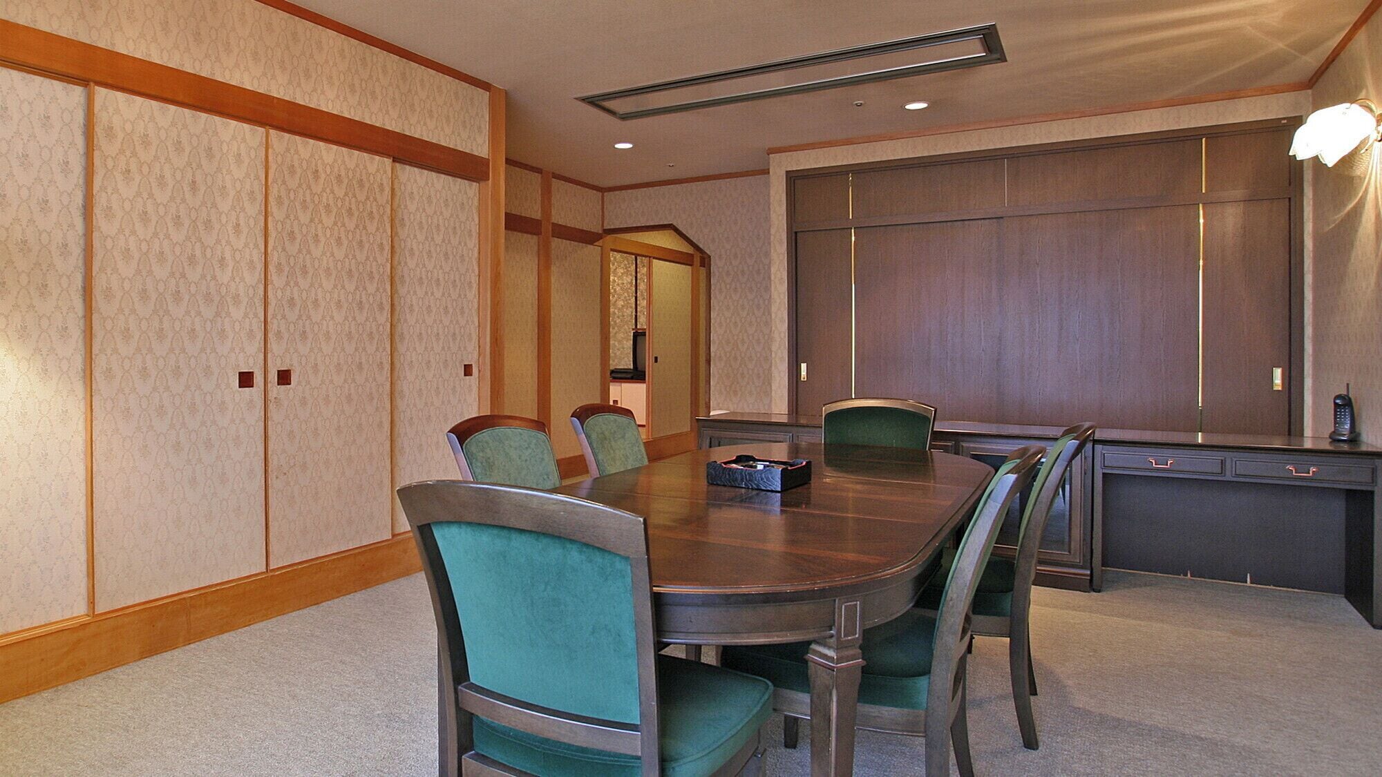 Une salle de conférence avec une table en bois, des chaises recouvertes d’un tissu vert, une petite table avec un téléphone et un grand miroir au mur.