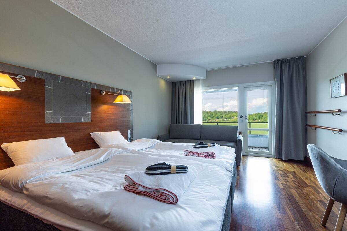 A hotel room with two beds, a sofa, a chair, a large window with a view of greenery, and a door leading to an outdoor area.