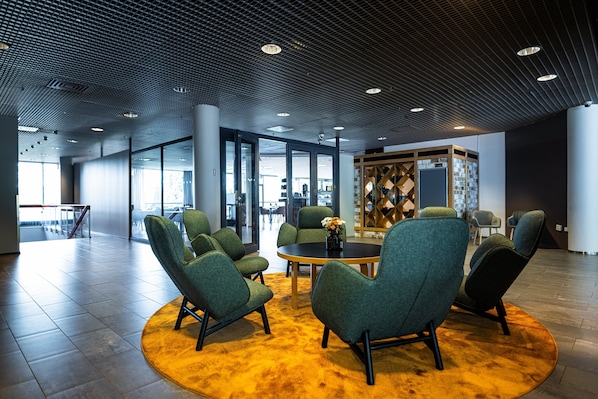 A modern lobby with green upholstered chairs around a central table, large windows, and a patterned ceiling.