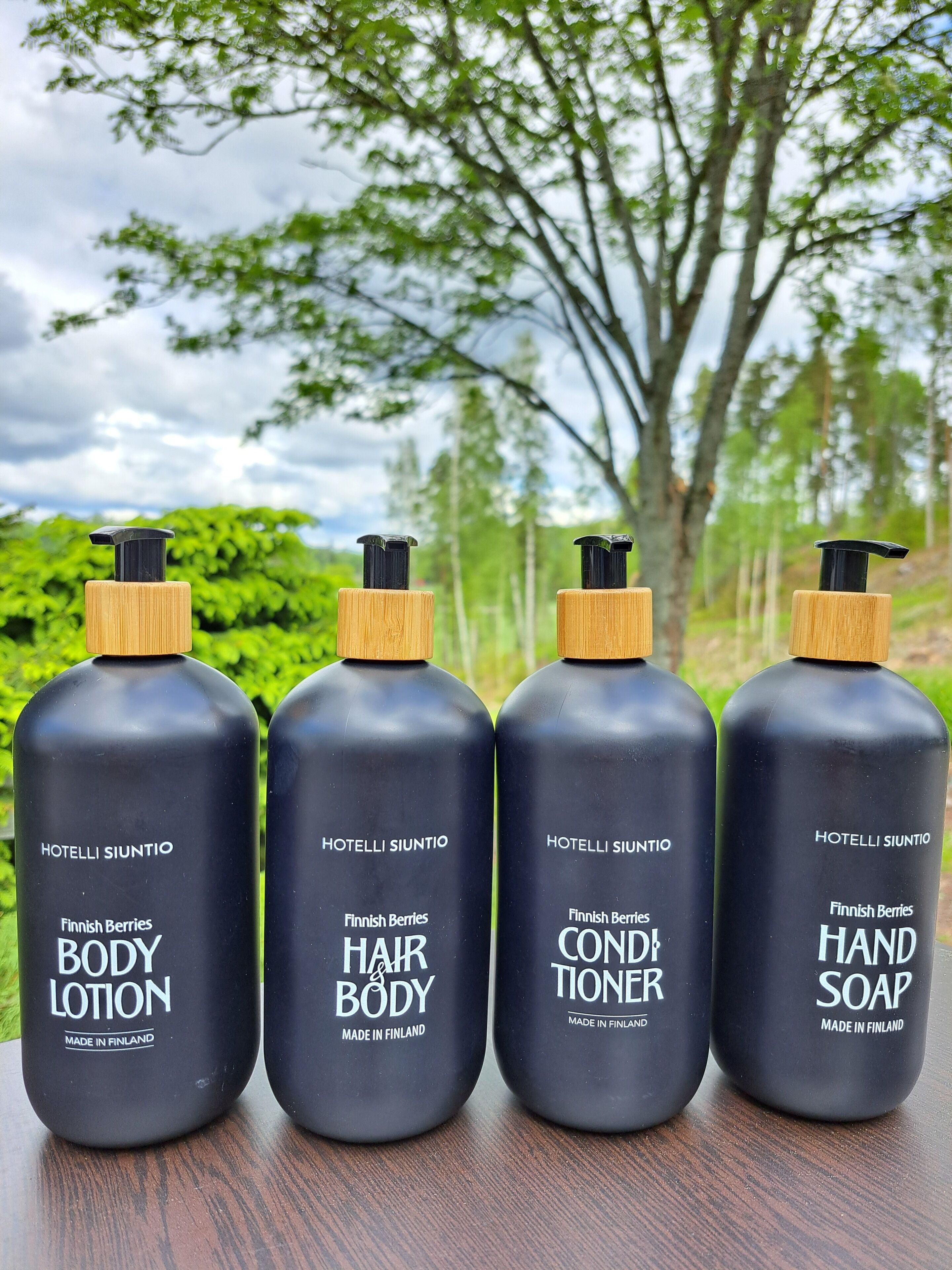 Quatre bouteilles de lotion corporelle, d’après-shampoing pour les cheveux et le corps, et de savon pour les mains, de fabrication finlandaise, posées sur une surface en bois, avec un arrière-plan naturel.