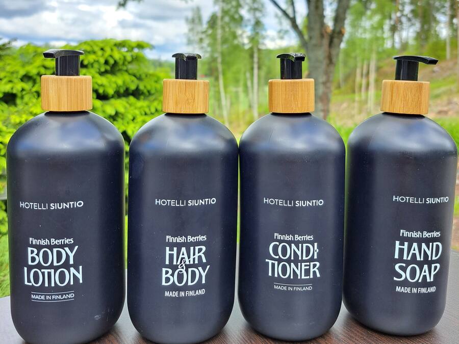 Quatre bouteilles de lotion corporelle, d’après-shampoing pour les cheveux et le corps, et de savon pour les mains, de fabrication finlandaise, posées sur une surface en bois, avec un arrière-plan naturel.
