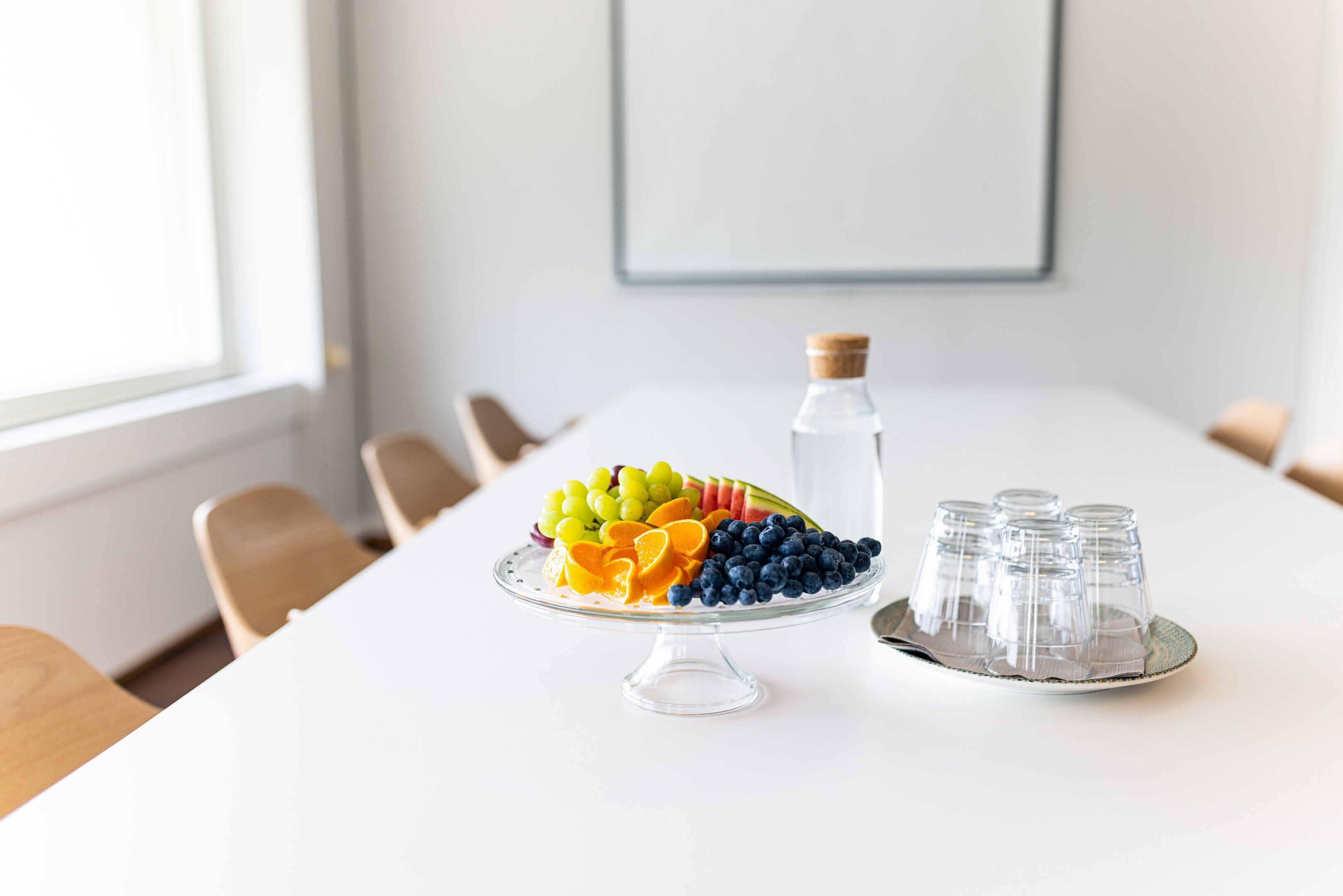 Una mesa de conferencias con una bandeja de frutas y vasos, una botella de agua y un cuadro en la pared.