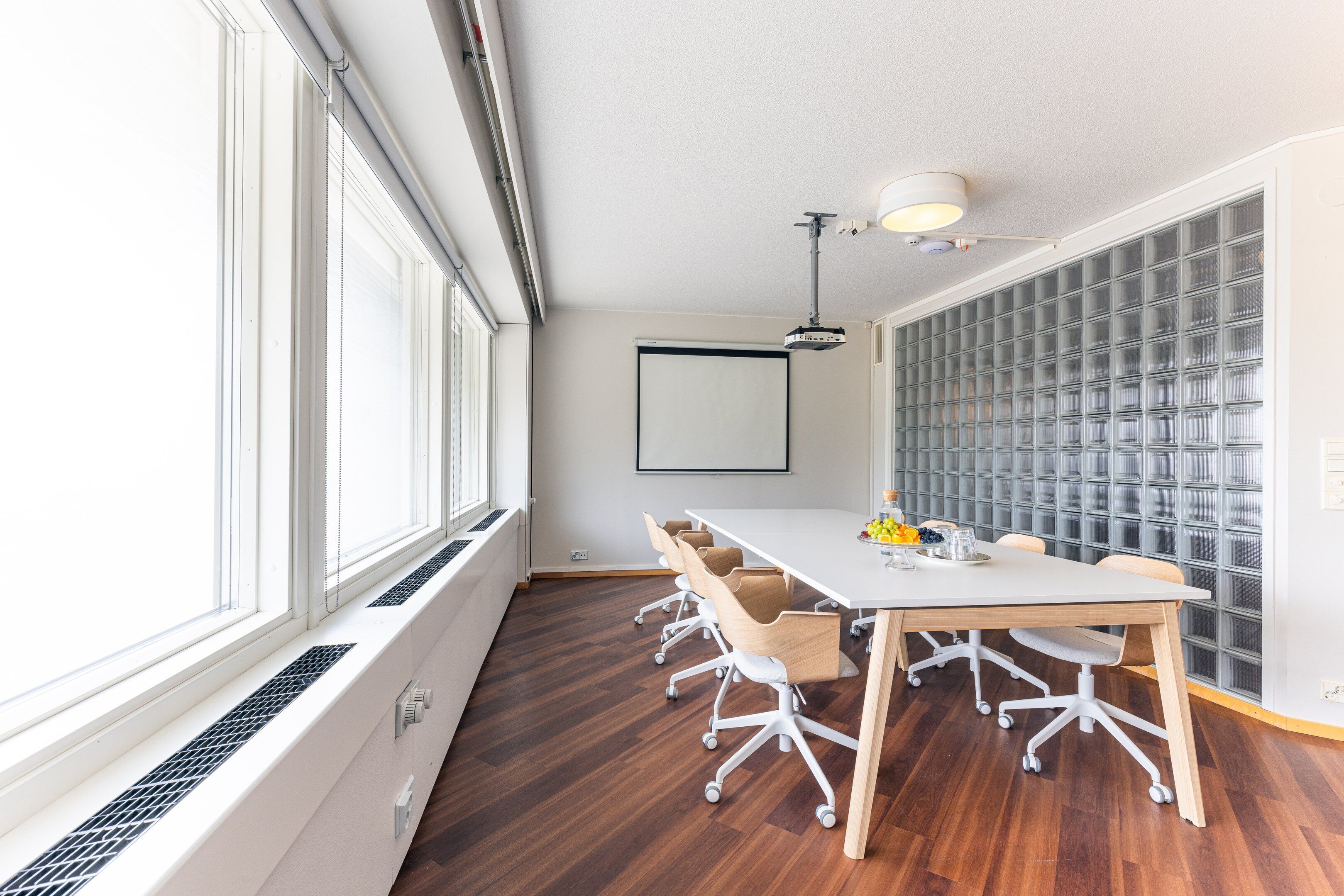 Un salón de conferencias con una mesa blanca, seis sillas, una pantalla de proyector y una ventana con persianas.