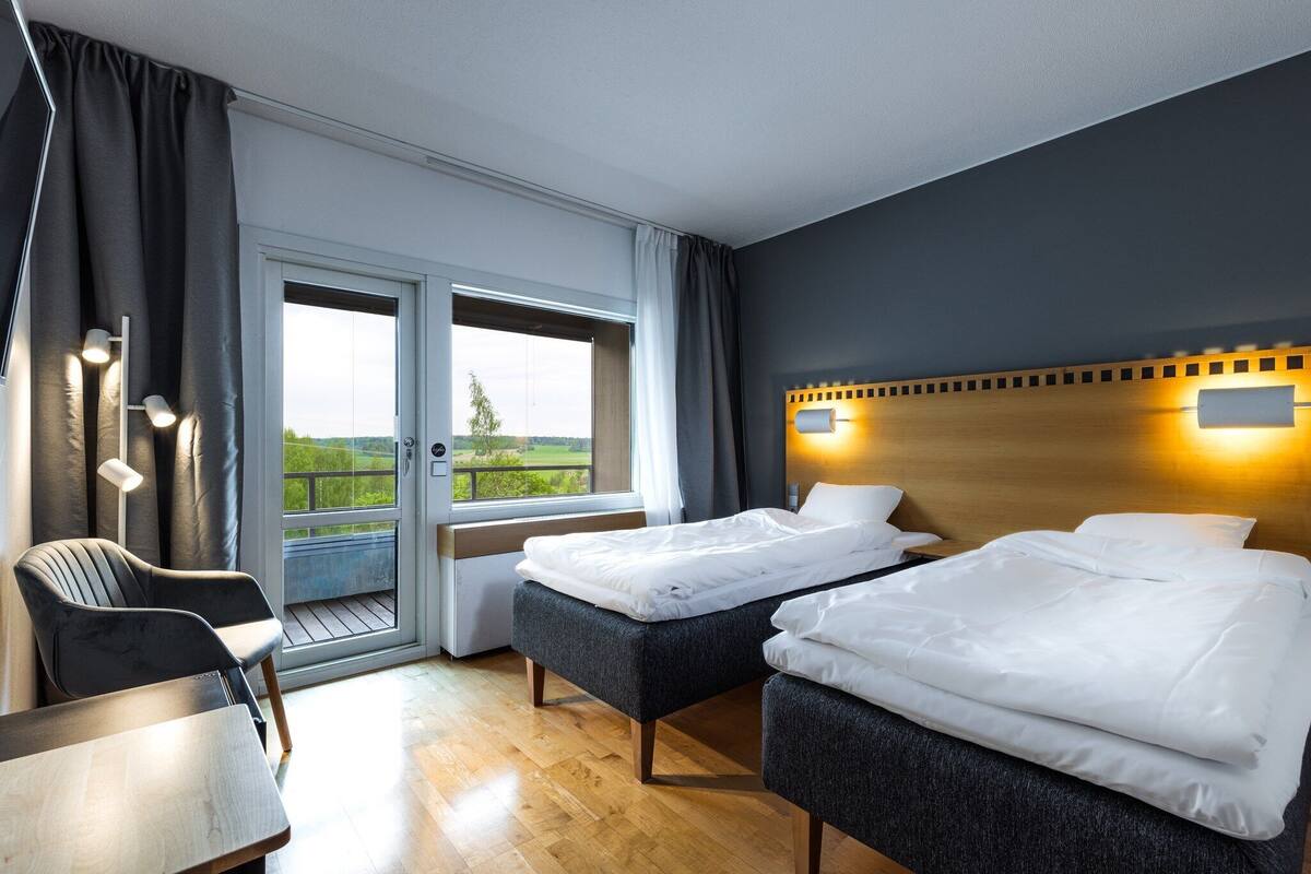 A hotel room with two beds, a desk, and a chair. There is a balcony with a view of greenery.