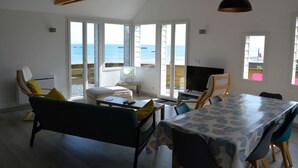A spacious living area with a large window overlooking the sea, a dining table, and a sofa. - Le Perchoir Avec vue Mer, à 300m de la Plage (Saint-Côme-de-Fresné)