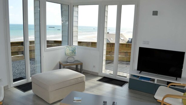 A modern living room with a large TV, a sofa, a coffee table, and a view of the beach through large windows. - Le Perchoir Avec vue Mer, à 300m de la Plage (Saint-Côme-de-Fresné)